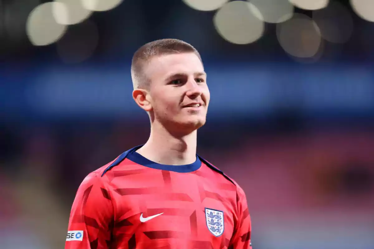 Un jugador de fútbol con una camiseta roja y el escudo de Inglaterra sonríe en un estadio iluminado. Un jugador de fútbol con una camiseta roja y el escudo de Inglaterra sonríe en un estadio iluminado.