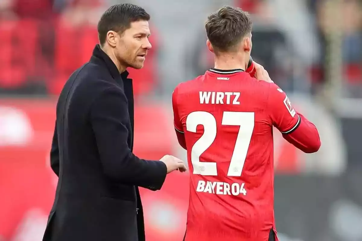 A soccer coach talks to a player wearing a red jersey with the number 27 and the name Wirtz. A soccer coach talks to a player wearing a red jersey with the number 27 and the name Wirtz.