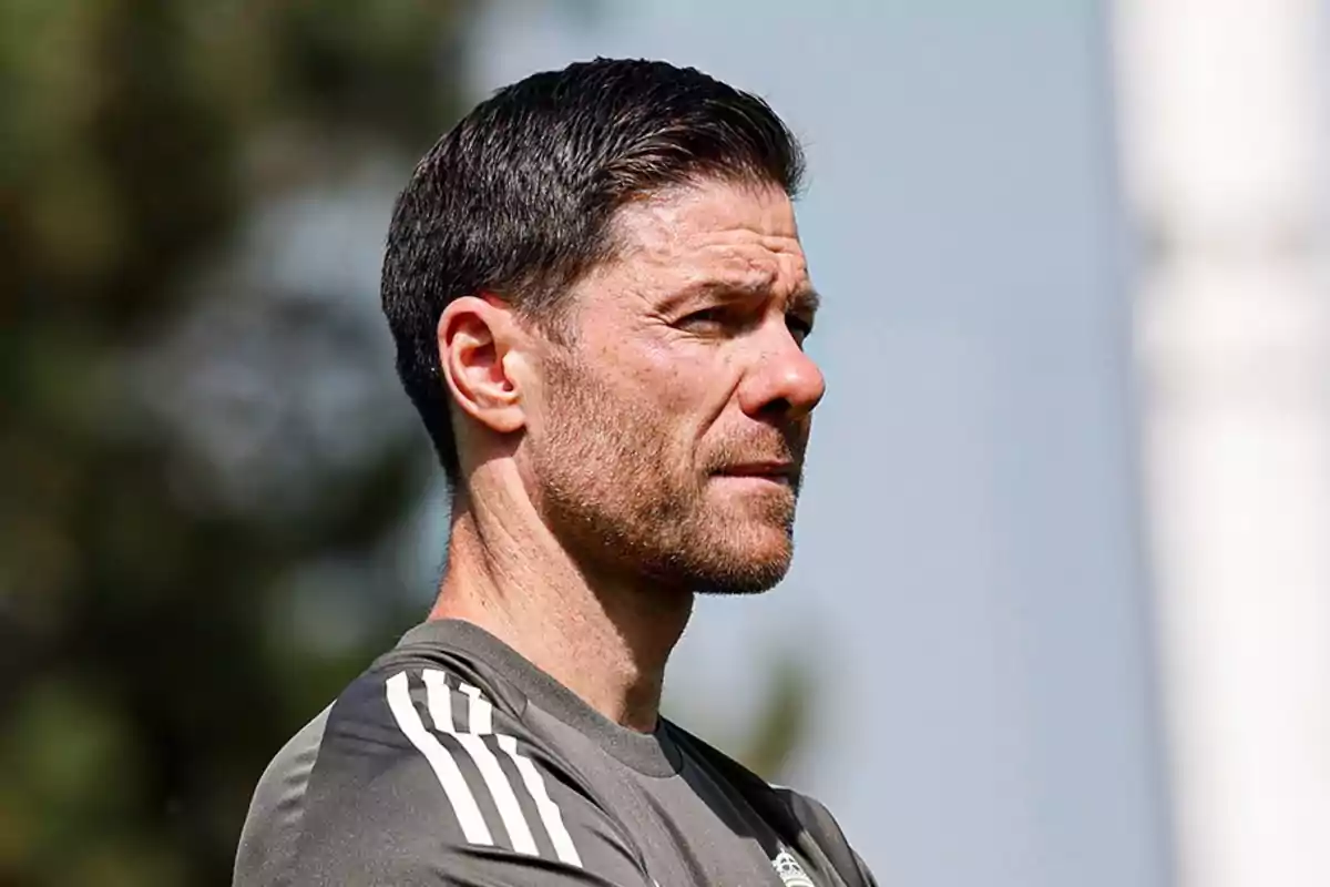 Hombre de cabello corto y barba mirando hacia la derecha con expresión seria, viste camiseta deportiva gris con rayas blancas en el hombro y fondo desenfocado al aire libre Hombre de cabello corto y barba mirando hacia la derecha con expresión seria, viste camiseta deportiva gris con rayas blancas en el hombro y fondo desenfocado al aire libre