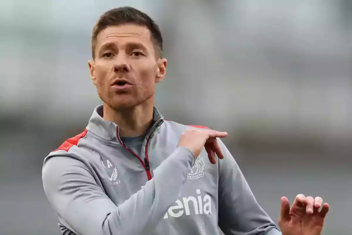 A man in a gray sports jacket with red details gesturing with his hand. A man in a gray sports jacket with red details gesturing with his hand.