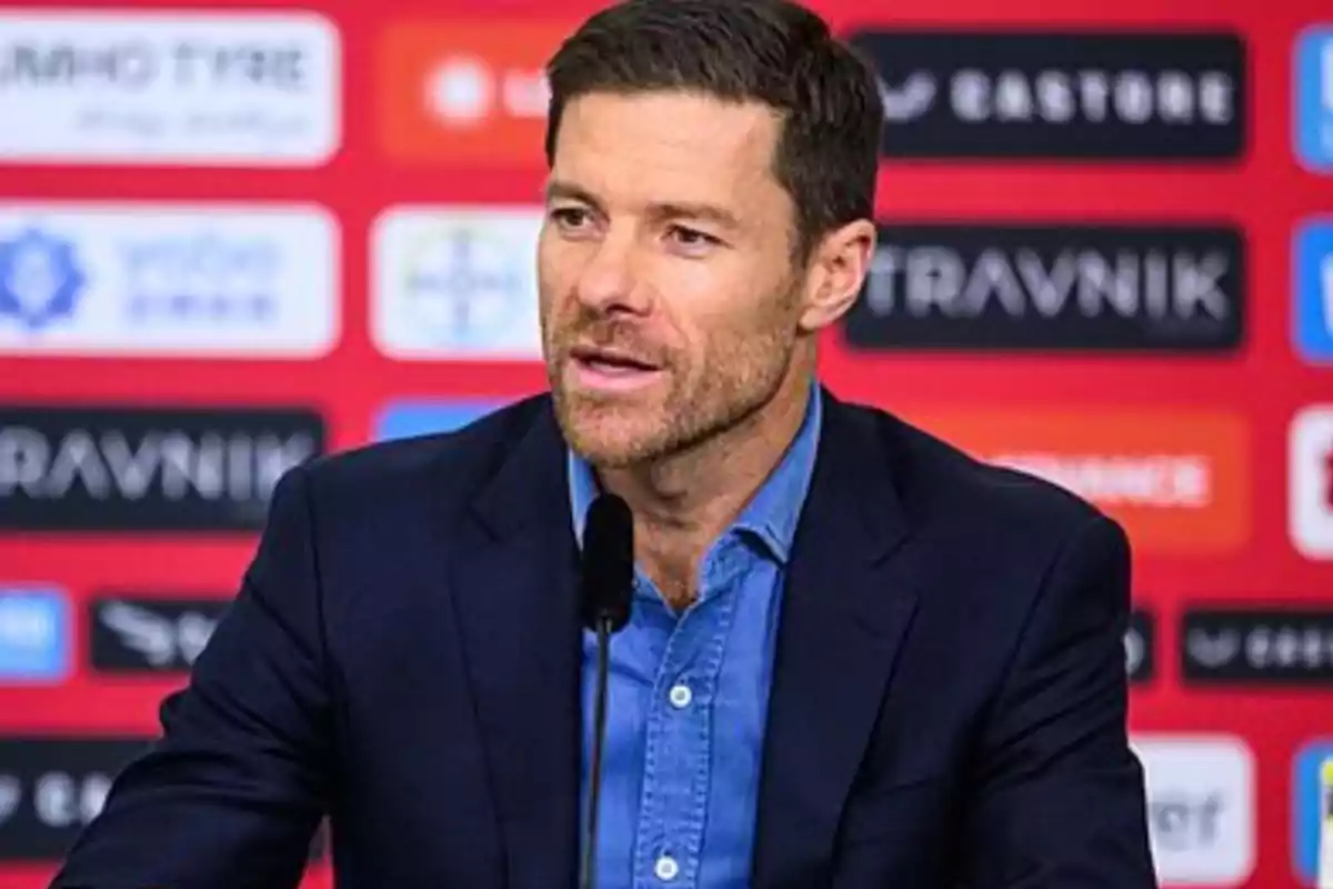 A man with a beard and short hair is speaking at a press conference with a red background full of logos. A man with a beard and short hair is speaking at a press conference with a red background full of logos.
