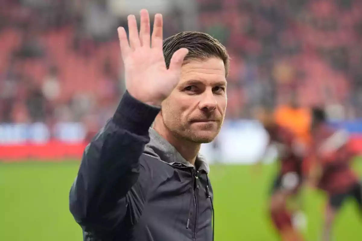 Un hombre con chaqueta oscura levanta la mano en un gesto de saludo en un estadio de fútbol. Un hombre con chaqueta oscura levanta la mano en un gesto de saludo en un estadio de fútbol.