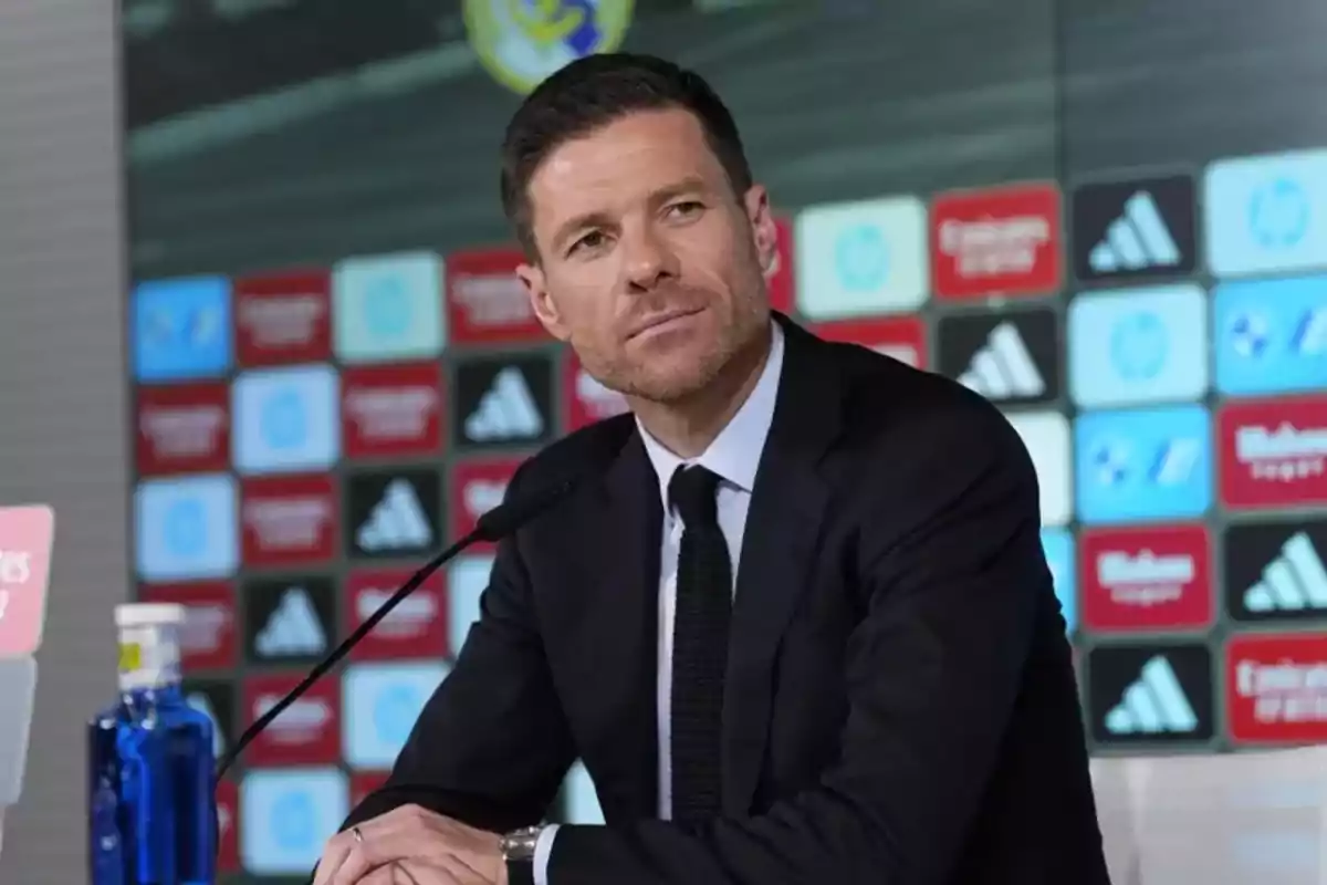 Man in a dark suit sitting in front of a microphone at a press conference with a background of sports logos Man in a dark suit sitting in front of a microphone at a press conference with a background of sports logos