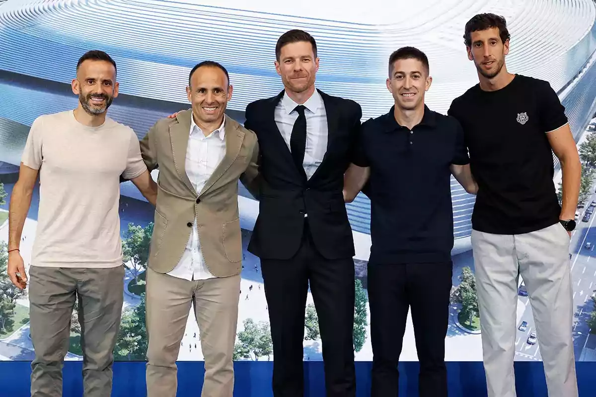 Cinco hombres posan juntos sonriendo frente a una imagen de un estadio moderno Cinco hombres posan juntos sonriendo frente a una imagen de un estadio moderno