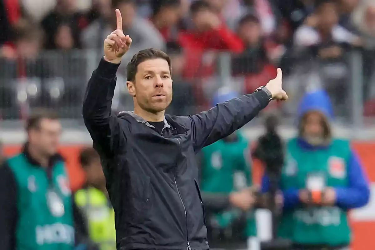 A man in a black jacket gestures with both hands at a sporting event.