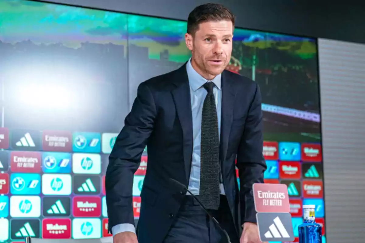 Hombre con traje oscuro y corbata hablando en una conferencia de prensa deportiva con fondo de patrocinadores y una botella de agua sobre la mesa Hombre con traje oscuro y corbata hablando en una conferencia de prensa deportiva con fondo de patrocinadores y una botella de agua sobre la mesa