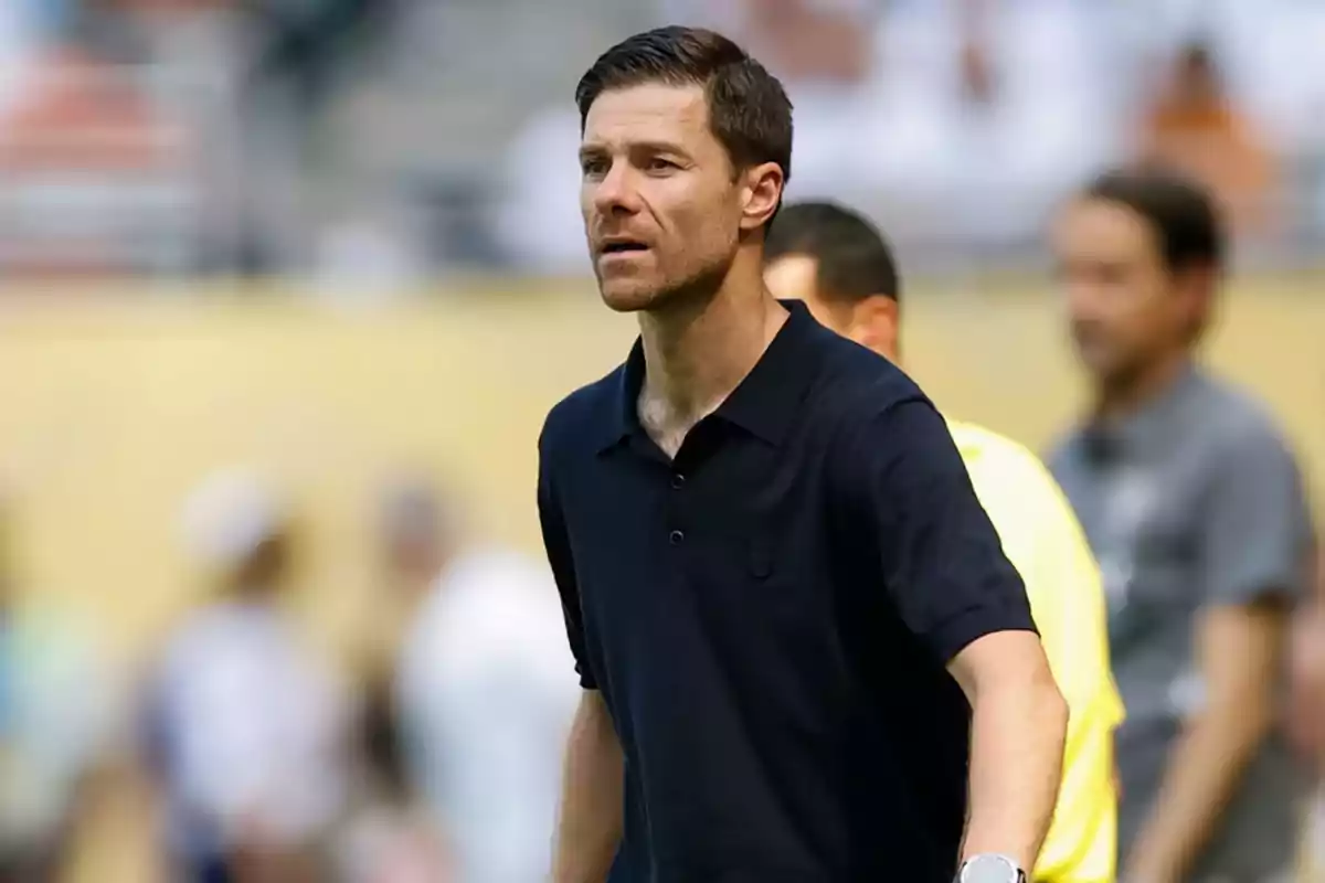 Short-haired man with a light beard wearing a dark polo jersey in a sports setting with blurred people in the background Short-haired man with a light beard wearing a dark polo jersey in a sports setting with blurred people in the background