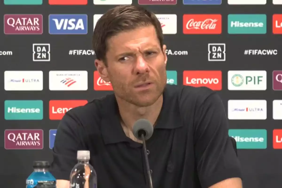 Man with a serious expression sitting in front of a microphone at a press conference with a backdrop of sponsor logos