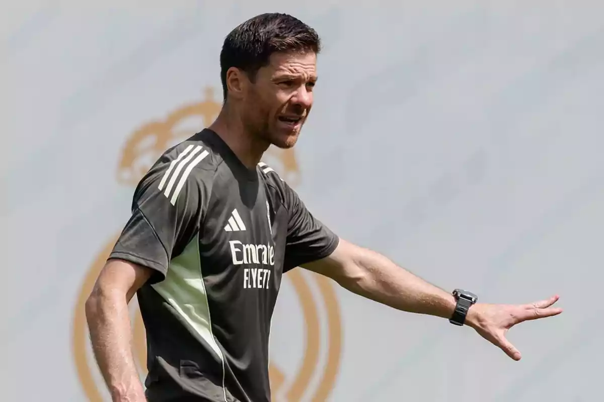 Entrenador de fútbol dando indicaciones en un campo de entrenamiento con camiseta deportiva oscura y fondo con el escudo de un club.