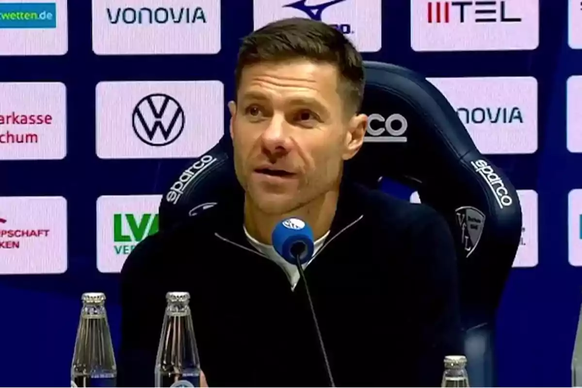 Un hombre en una conferencia de prensa sentado frente a un micrófono con logotipos de patrocinadores en el fondo. Un hombre en una conferencia de prensa sentado frente a un micrófono con logotipos de patrocinadores en el fondo.