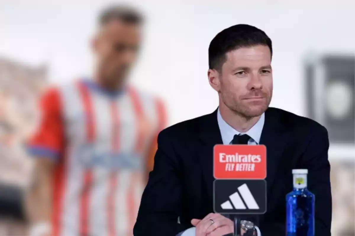 Man in a suit at a press conference with an Emirates microphone and a bottle of water, in the background a blurred figure wearing a red and white striped jersey