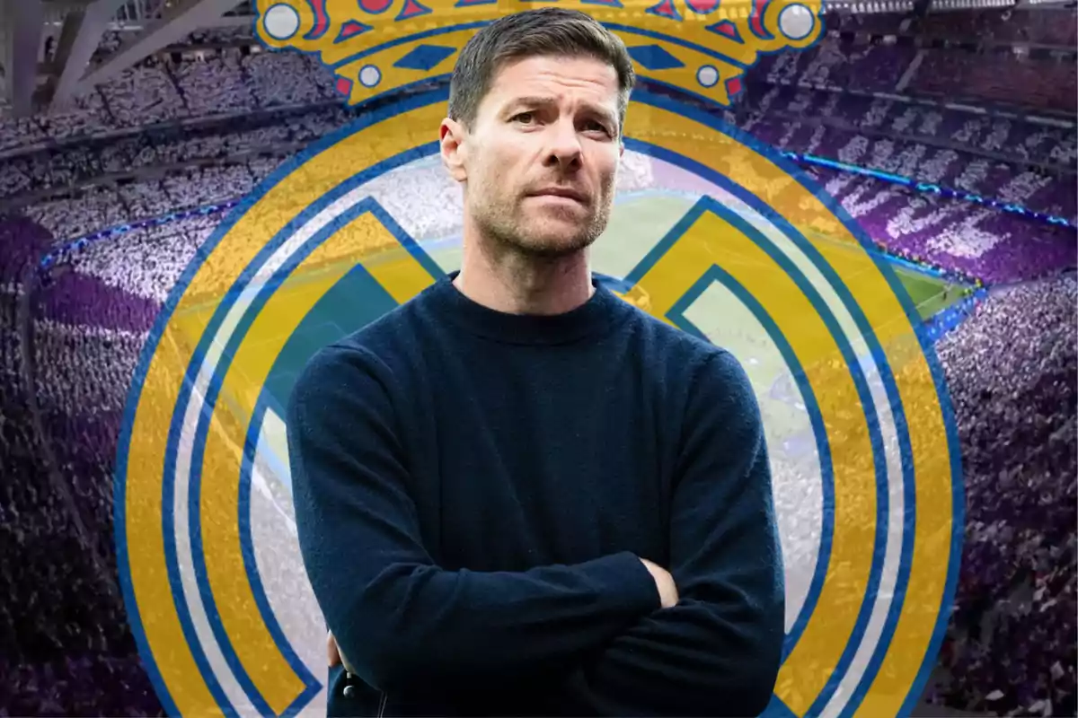 A man in a dark sweater poses with his arms crossed in front of a full stadium and a soccer emblem in the background.