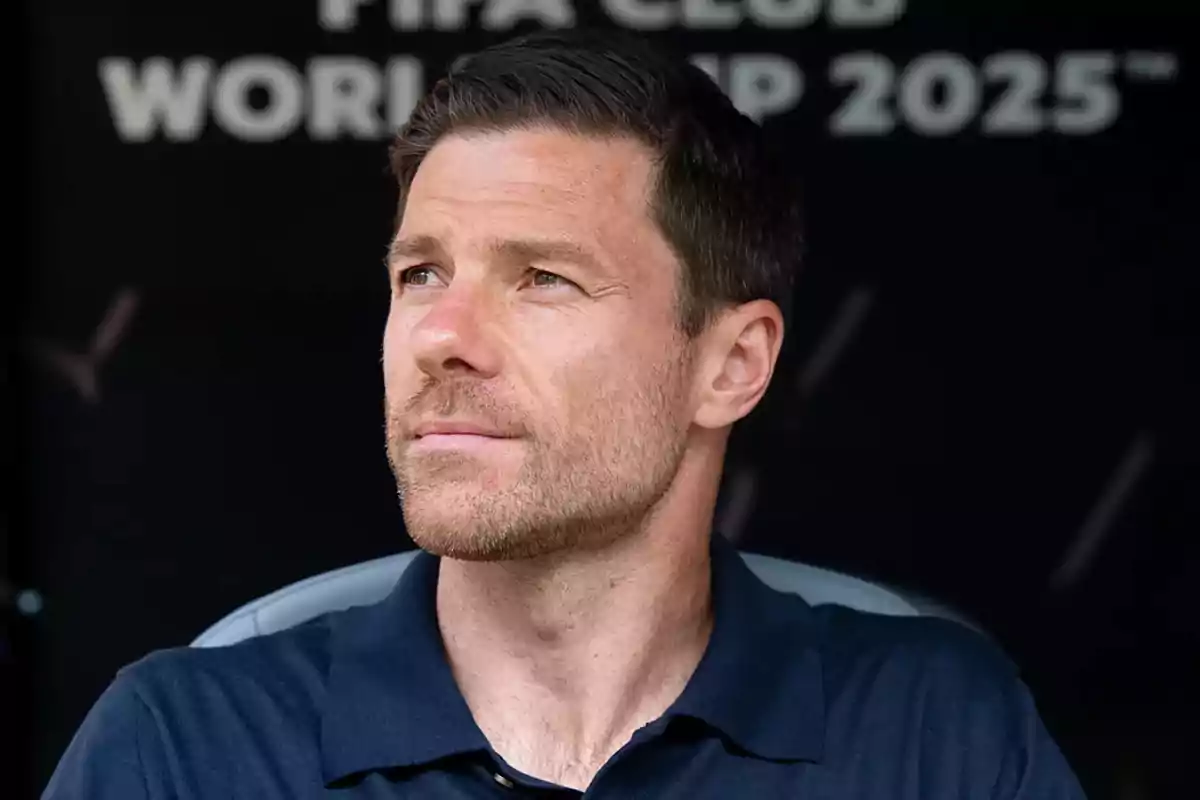 Hombre de cabello corto y barba ligera con camisa azul marino mirando hacia un lado con expresión seria y fondo oscuro con letras desenfocadas. Hombre de cabello corto y barba ligera con camisa azul marino mirando hacia un lado con expresión seria y fondo oscuro con letras desenfocadas.