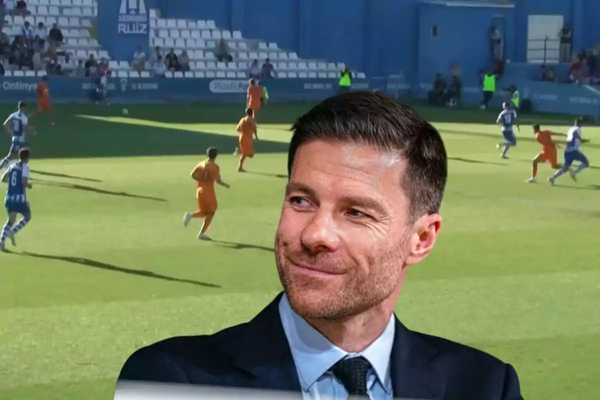 Hombre con traje sonriente en primer plano con un partido de fútbol de fondo en un estadio con gradas blancas y jugadores en uniforme naranja y azul