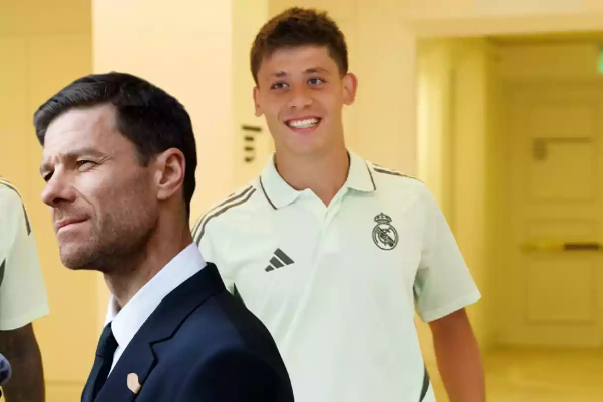 Two men, one in a suit and the other in a Real Madrid jersey, pose in a brightly lit hallway.