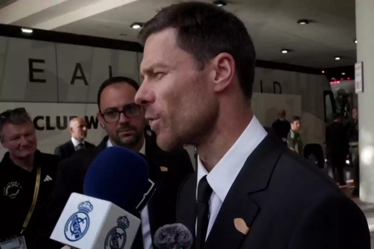 A man in a dark suit is being interviewed by the press in front of a bus with the Real Madrid logo while several people watch in the background.