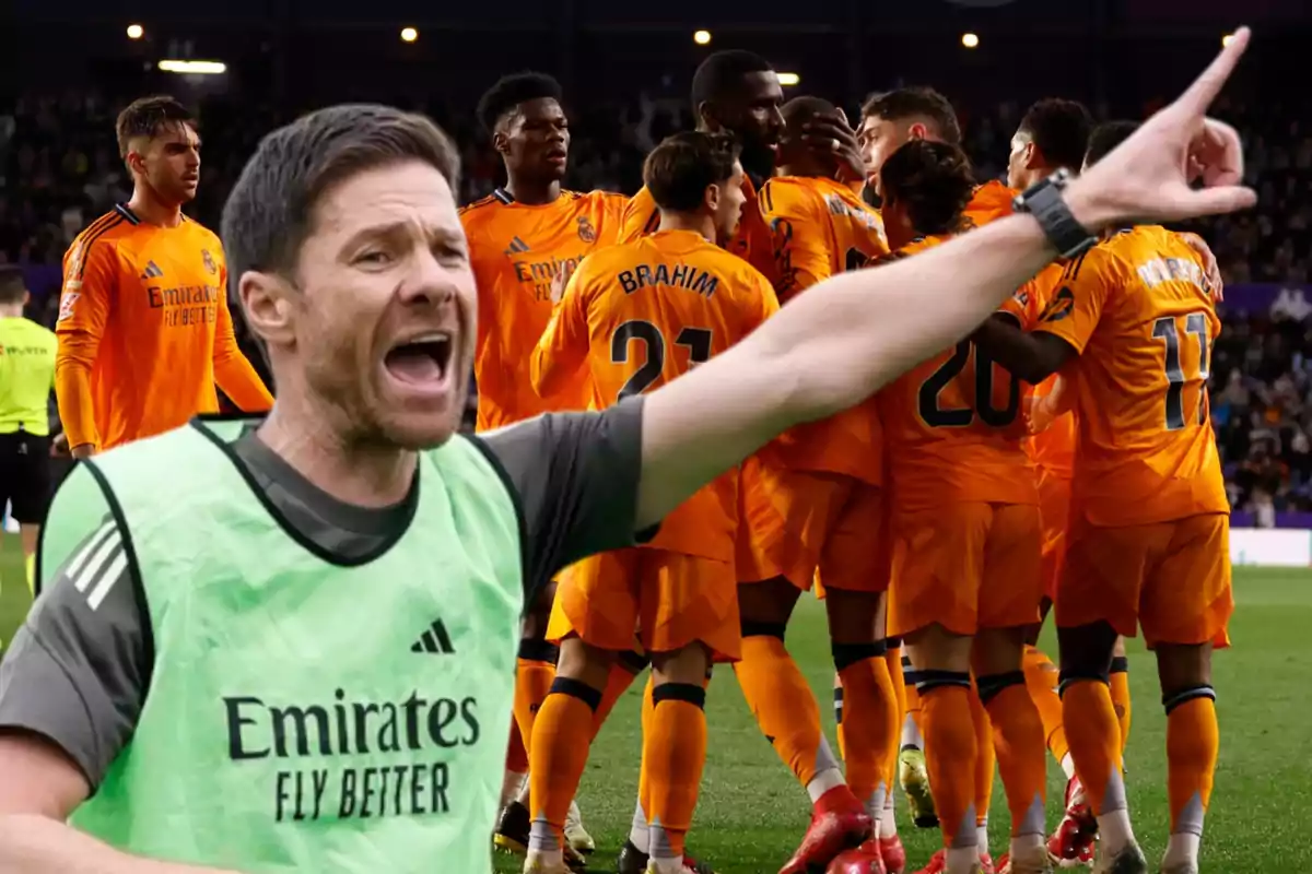 Un entrenador de fútbol da indicaciones en primer plano mientras un grupo de jugadores con uniforme naranja celebra en el campo