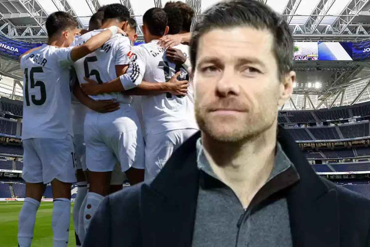 A group of players hug on the field while a man in a dark jacket appears in the foreground inside a stadium.