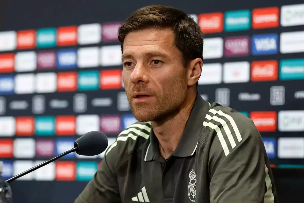 Man wearing a Real Madrid jersey speaking at a press conference in front of a microphone and a sponsor backdrop. Man wearing a Real Madrid jersey speaking at a press conference in front of a microphone and a sponsor backdrop.
