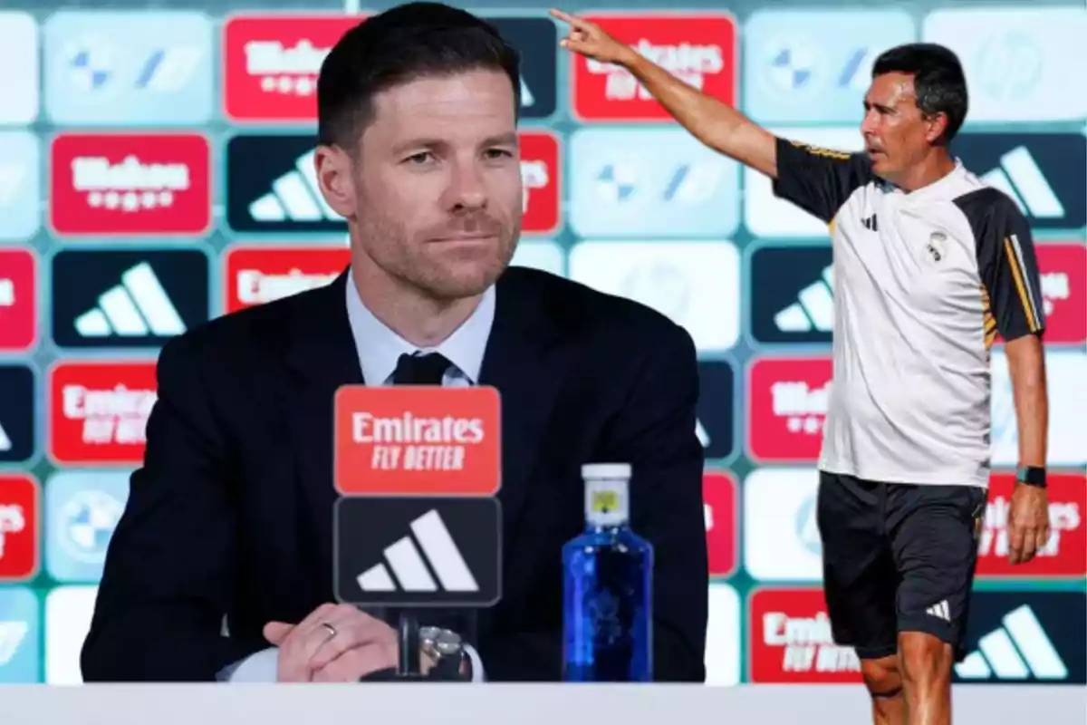 A man in a suit is sitting at a press conference while another man in sportswear points forward, both with a backdrop of sports sponsors.