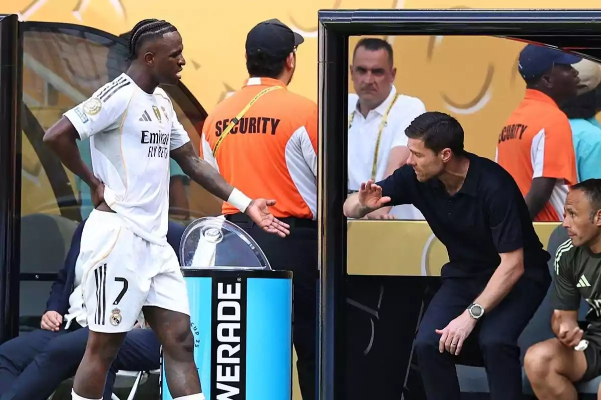 Un jugador de fútbol del Real Madrid discute con un entrenador cerca del área técnica mientras personal de seguridad observa en el fondo
