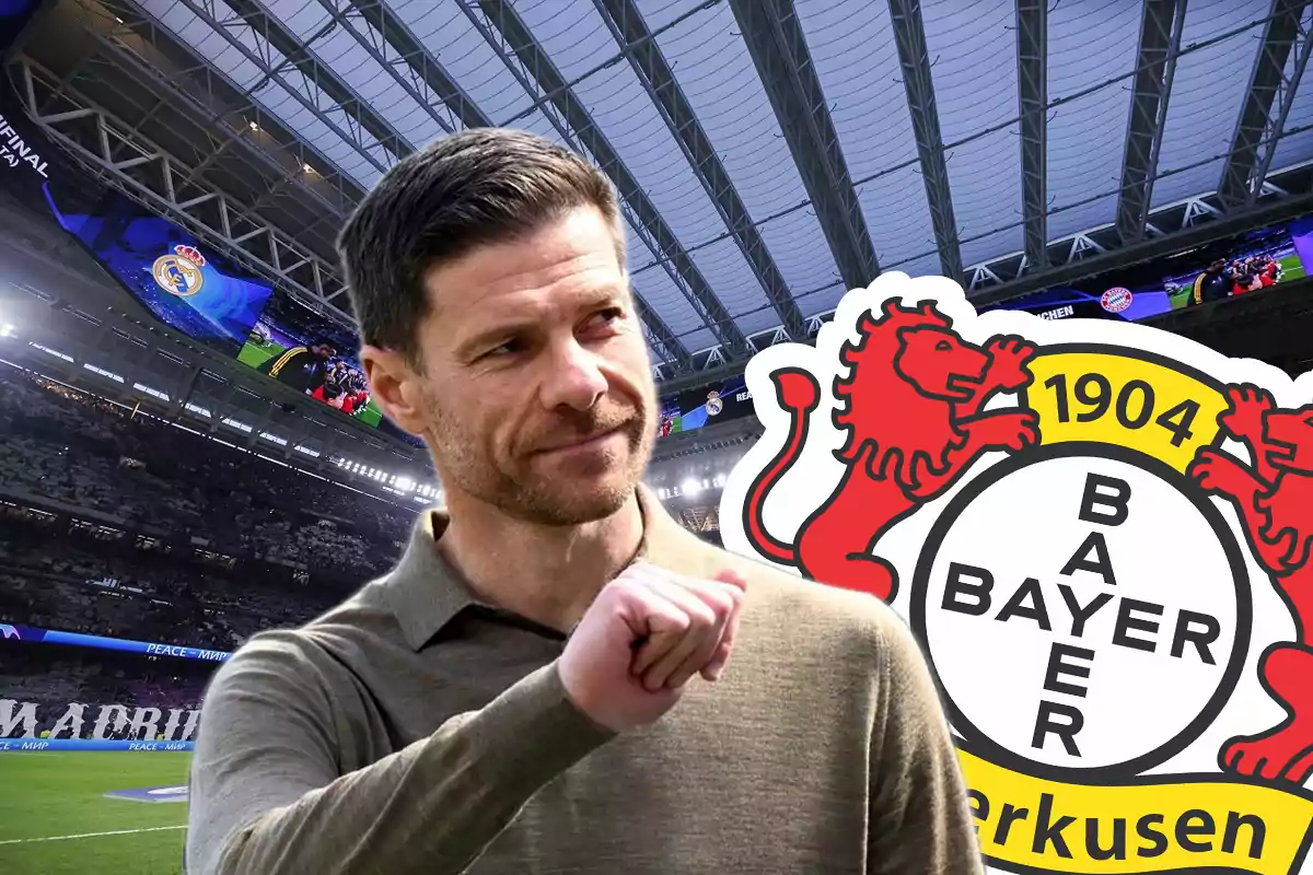 Un hombre con barba y cabello corto viste una camisa beige y aparece en primer plano con el logo del Bayer Leverkusen a su lado y el interior de un estadio de fútbol de fondo