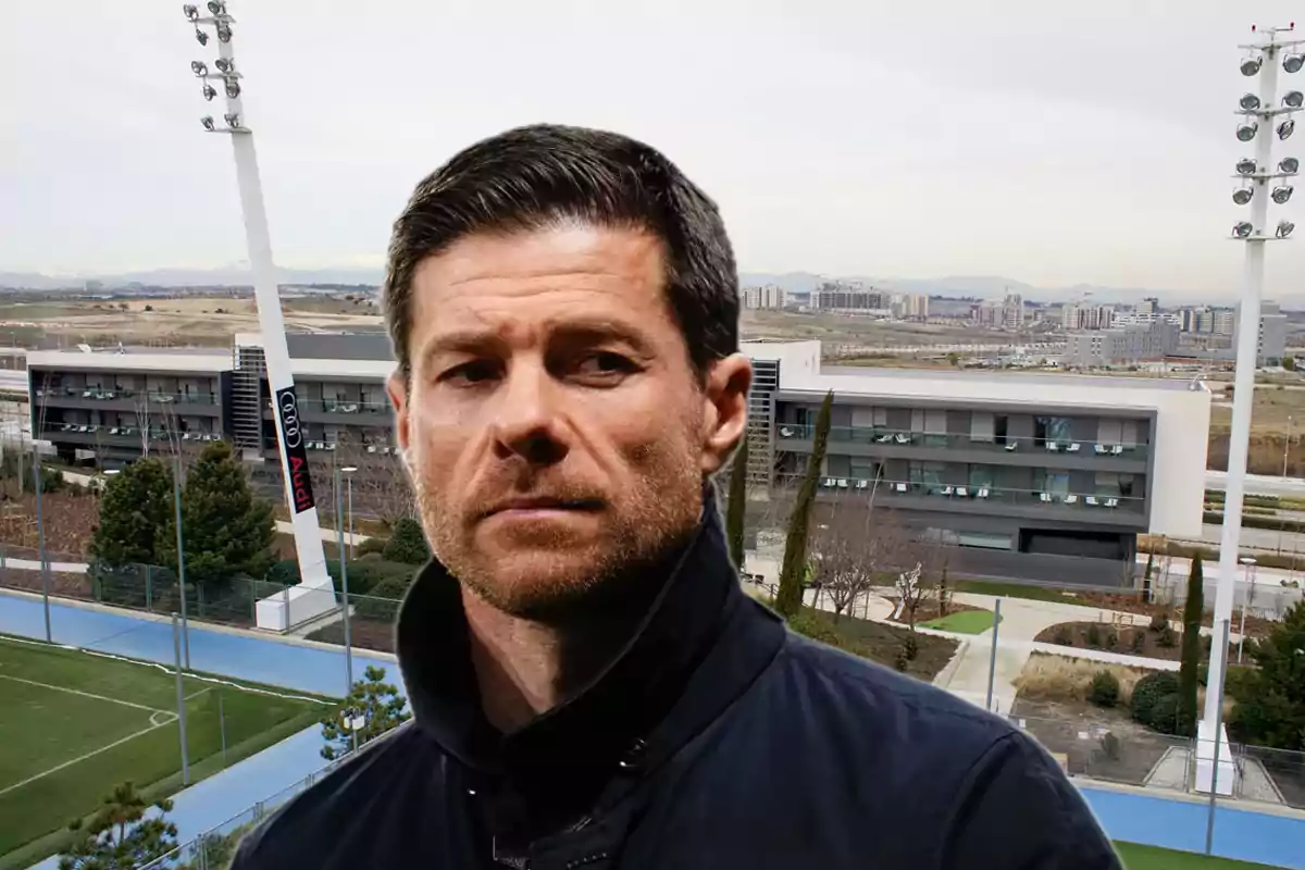 Short-haired man with a trimmed beard and a serious expression in front of a modern sports complex with soccer fields and buildings in the background