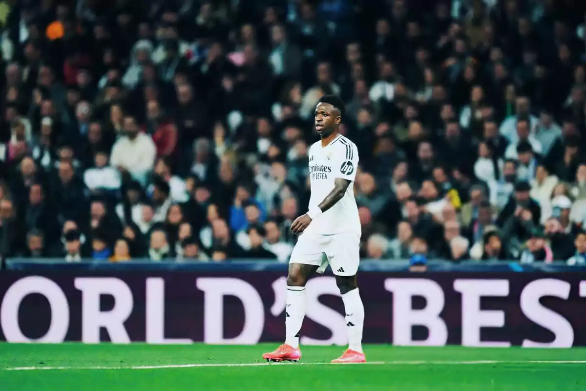 Un jugador de fútbol con uniforme blanco camina en el campo durante un partido, con una multitud de espectadores en el fondo.