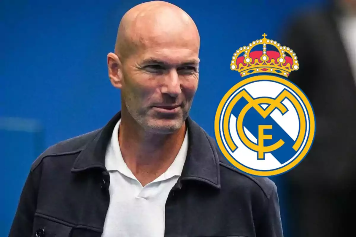 Un hombre calvo con chaqueta oscura y camisa blanca está de pie junto a un logotipo del Real Madrid sobre un fondo azul.