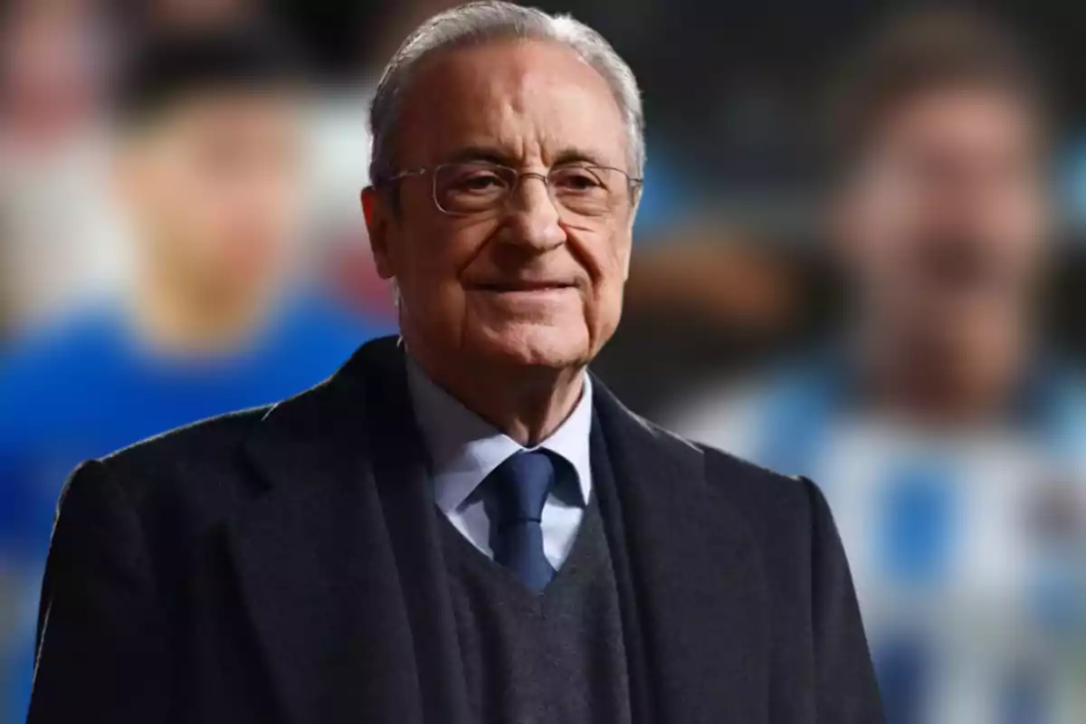 An older man with glasses and a dark suit slightly smiles while standing in front of a blurred background.