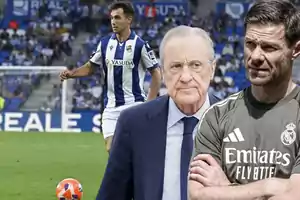 A Real Sociedad player on the field with two men in the foreground, one in a suit and the other in a Real Madrid training jersey.
