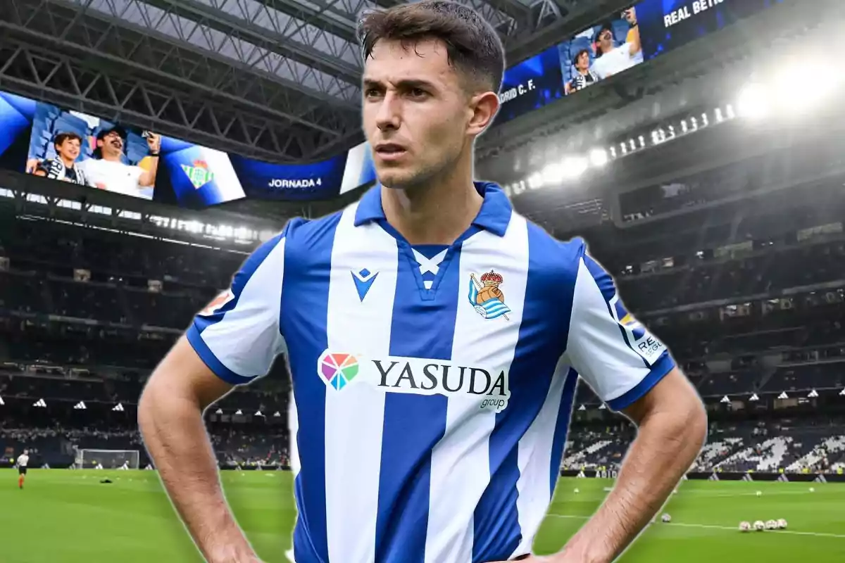Soccer player wearing a Real Sociedad uniform posing in a football stadium full of lights and screens.